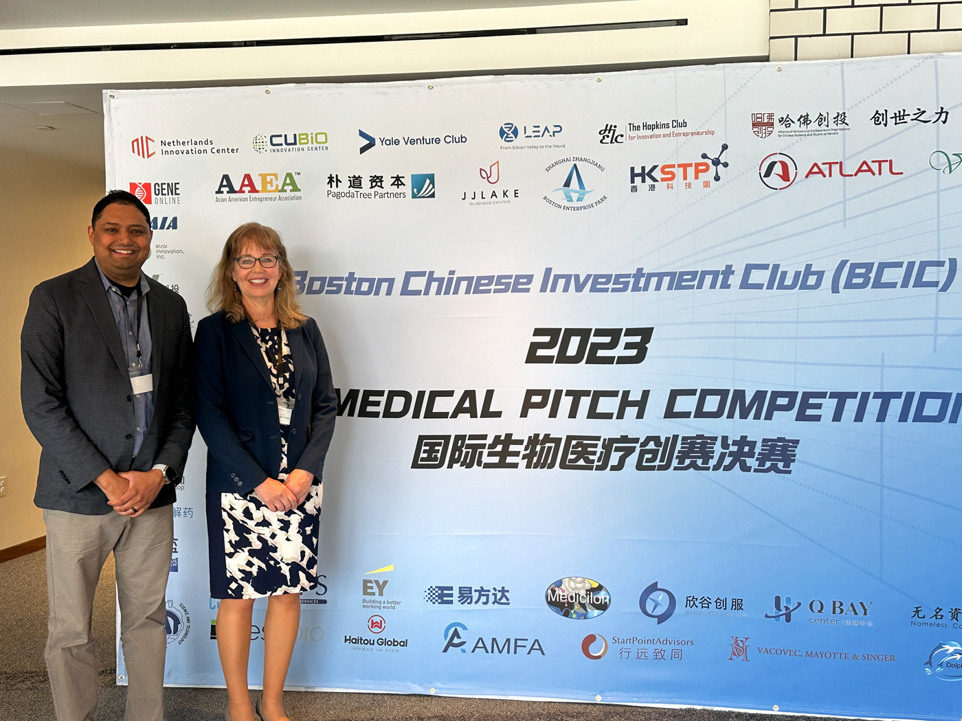 two of Likarda's representatives, a man and a woman, stand in front of the boston chinese investment club 2023 medical pitch competition photo background. On the background are also sponsors of the event.